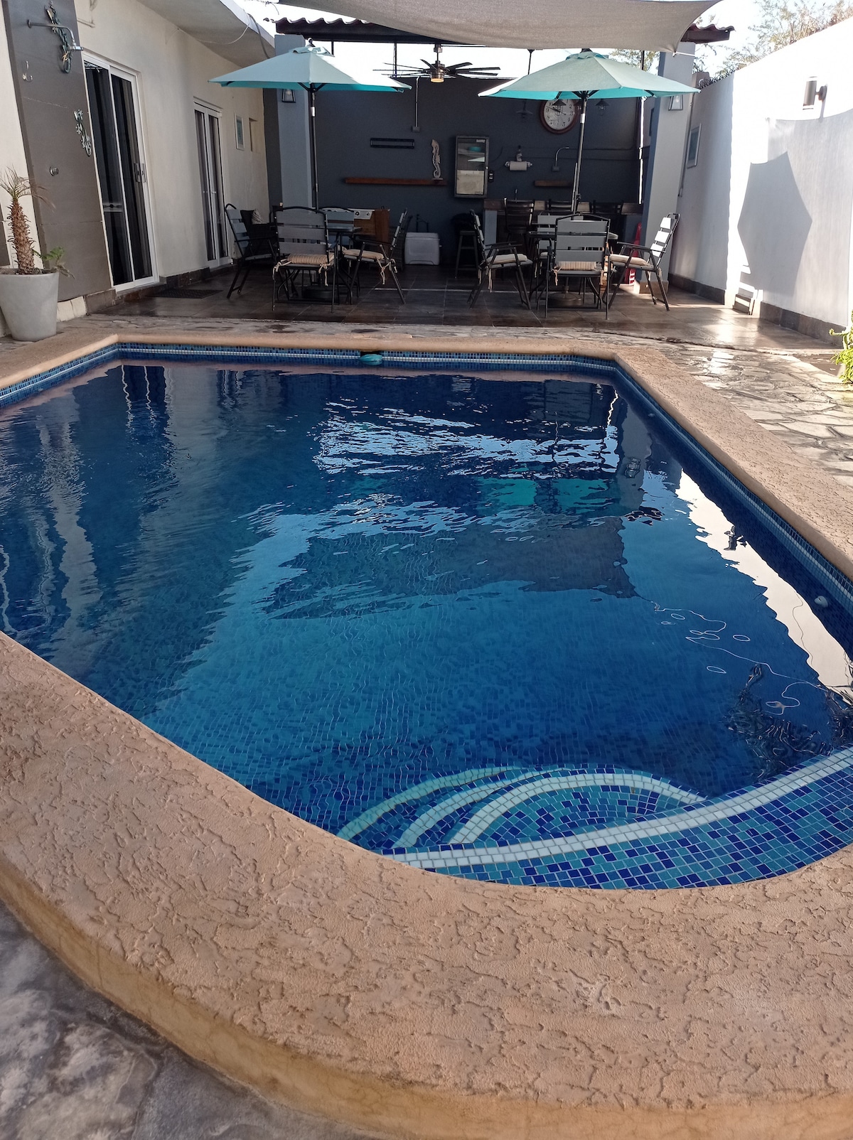 A small pool is framed by a textured stone edge, with clear blue water reflecting sunlight. Dark patio furniture can be seen in the background, accompanied by green umbrellas. The area is designed for relaxation and outdoor dining.