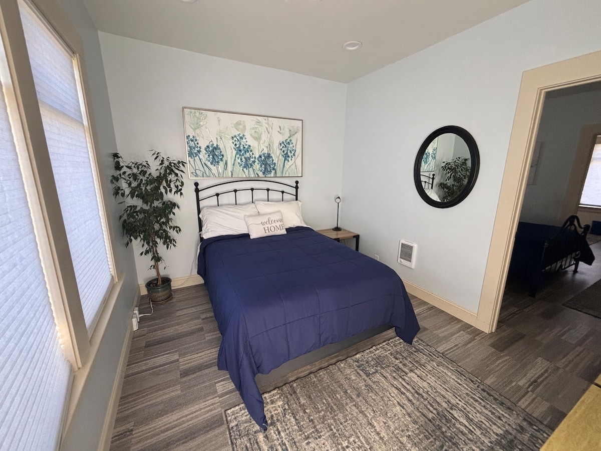 A bright bedroom features a spacious bed dressed in dark bedding. A decorative wall piece with botanical prints complements the room. Natural light filters through the partially open blinds, and a mirror hangs on one wall. A small plant adds a touch of greenery.