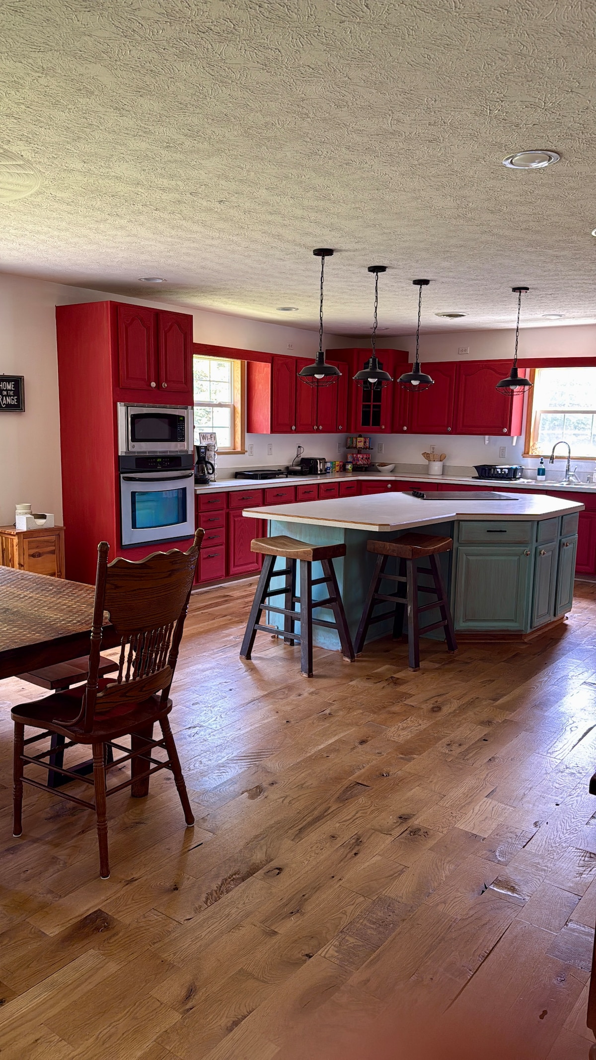 A spacious kitchen features bright red cabinetry and a central teal island with seating. Wood flooring is complemented by natural light streaming through windows. An adjacent dining area includes a wooden table and chairs, creating a welcoming space for meal preparation and gatherings.