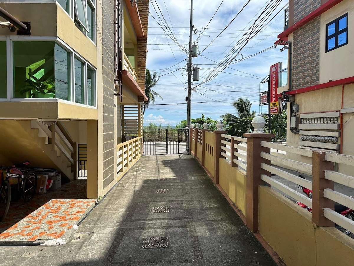 A pathway leads between two apartment buildings, bordered by a decorative fence. Electric lines are visible overhead, and a clear view of the sky can be seen in the distance. The ground is paved, with bicycles parked to one side.
