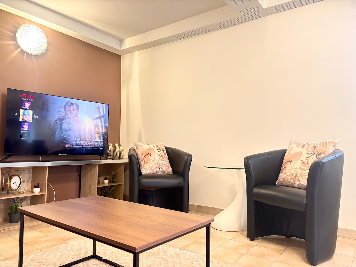 A cozy living area is presented, featuring two black armchairs with decorative cushions. A modern glass-topped table sits in front of them. A flat-screen TV is mounted on the wall, displaying a streaming service interface. Warm lighting enhances the inviting feel of the space.