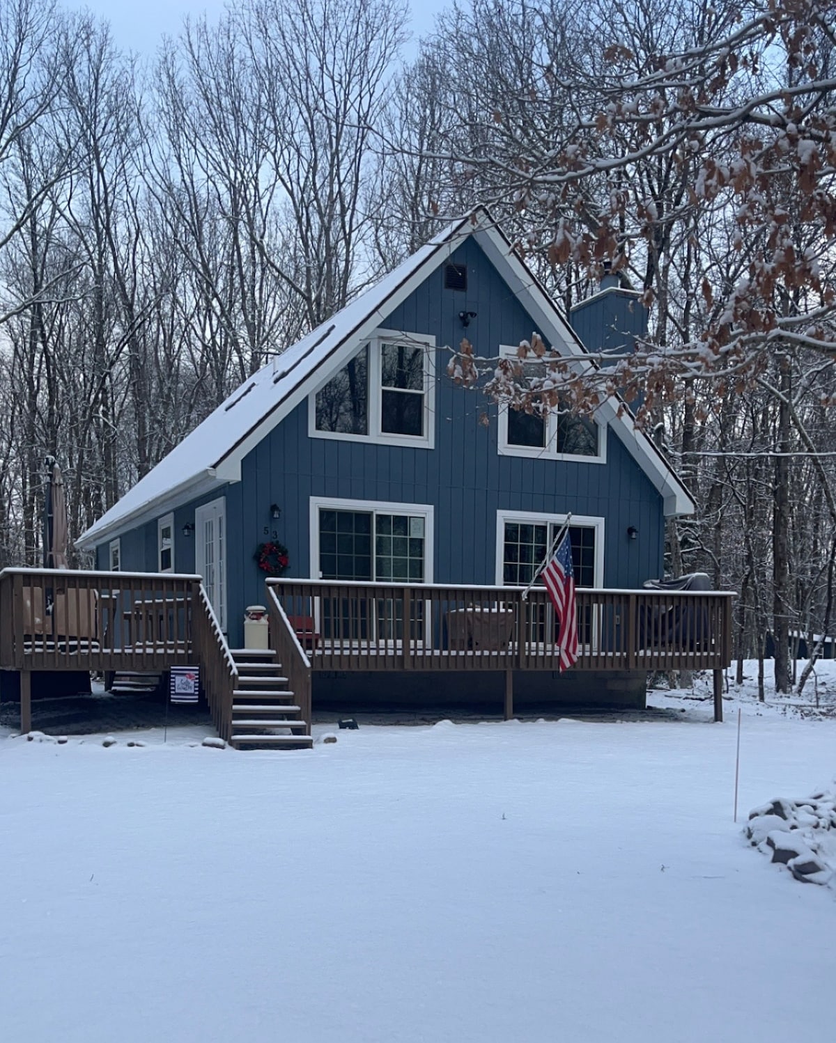 A charming blue house is surrounded by snow-covered trees. A welcoming porch is highlighted by a wooden railing, and an American flag is displayed. Large windows invite natural light, while the roof gently slopes, adding to the building's aesthetic. Snow blankets the ground, enhancing the serene atmosphere.