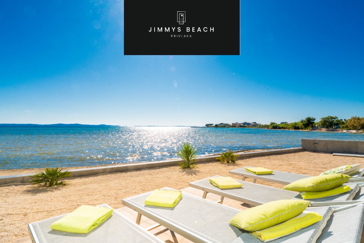 A serene beachfront is depicted with several sun loungers positioned on a sandy area. The clear blue water of the sea sparkles under the sunlight, with a distant view of the shoreline and a few trees, creating a relaxing environment.