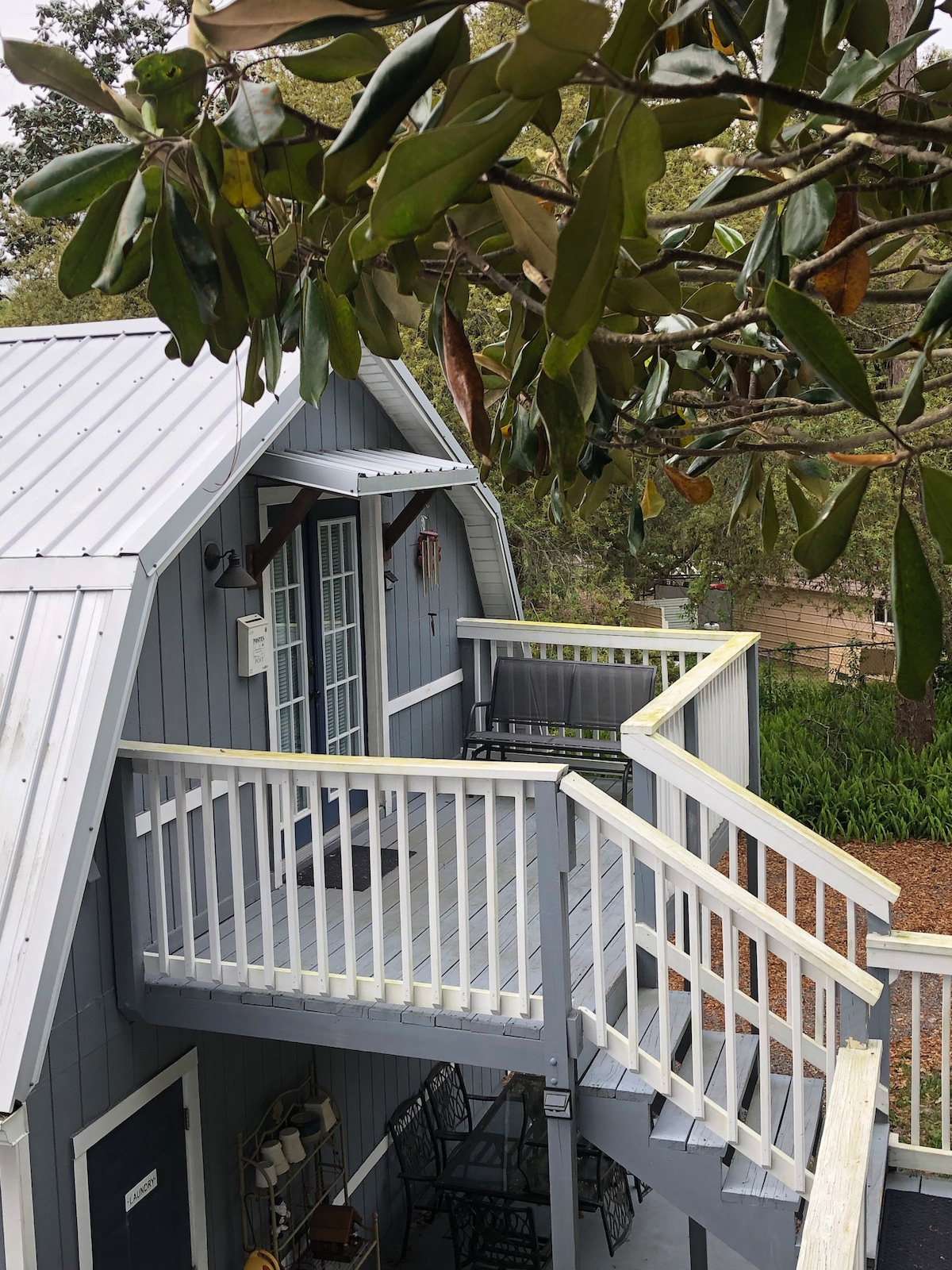 A second-story balcony features white railings and overlooks a lush yard. Two chairs are positioned on the balcony, while the building's grey exterior is complemented by a metal roof. Leafy branches frame the upper section of the image.
