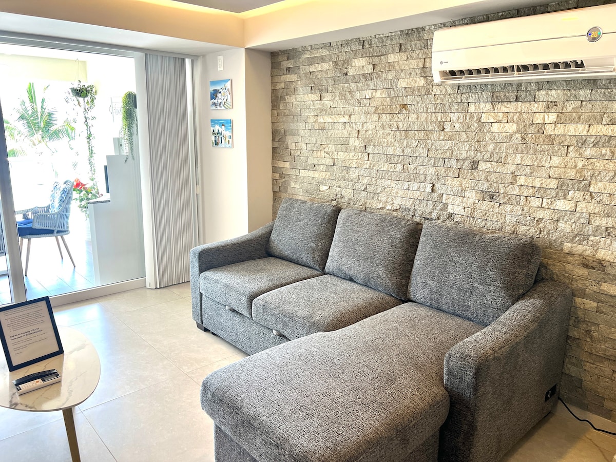 A spacious living area features a large gray sectional sofa, complemented by a natural stone accent wall. Expansive glass sliding doors offer views of the outdoor space. A round table sits nearby, emphasizing an open and bright atmosphere.