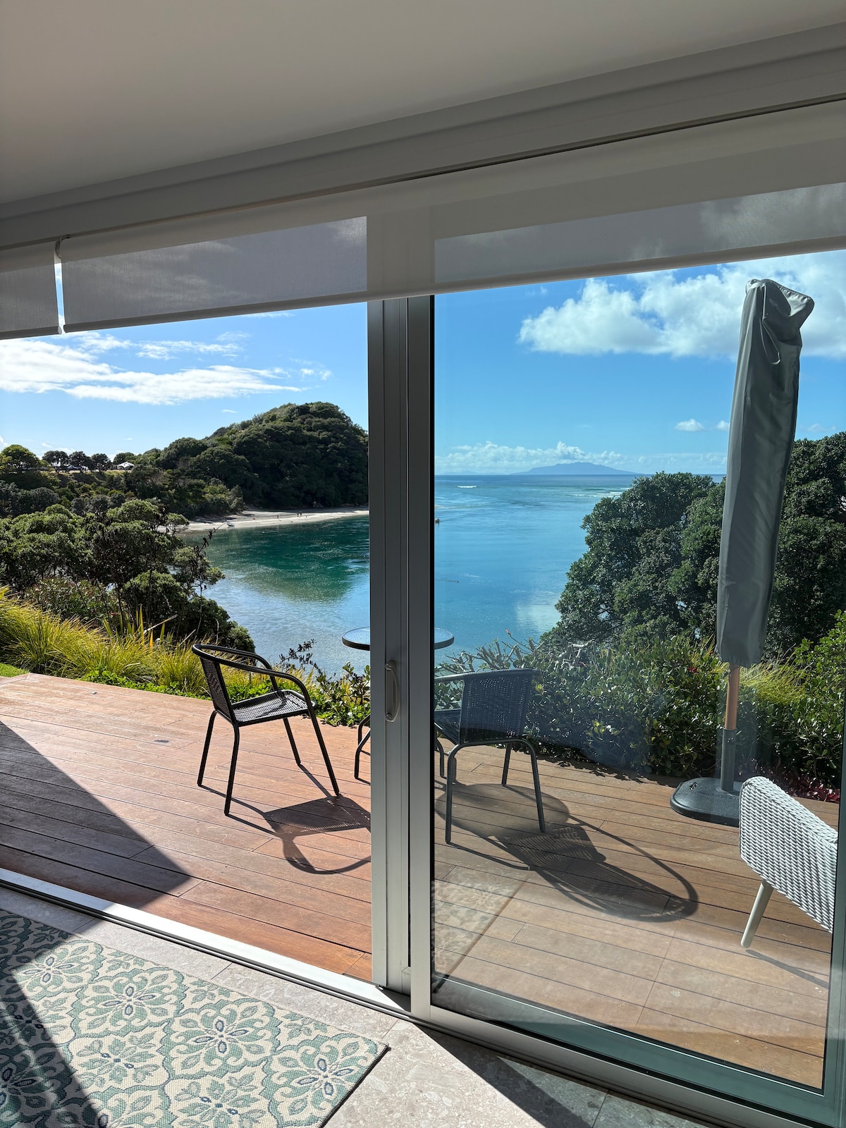 A sliding glass door leads to a private deck featuring two chairs and a small table, offering views of a serene bay and lush greenery. Natural light fills the indoor space, complemented by the tranquil blue water and distant shoreline visible beyond.