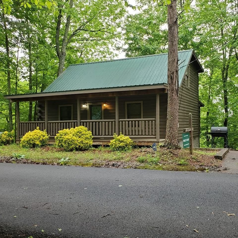 A two-story log cabin, surrounded by lush greenery. A covered front porch with wooden railing provides access, and colorful shrubs are planted in front. The metal roof is painted green, blending with the natural landscape. A charcoal grill is positioned to the side.