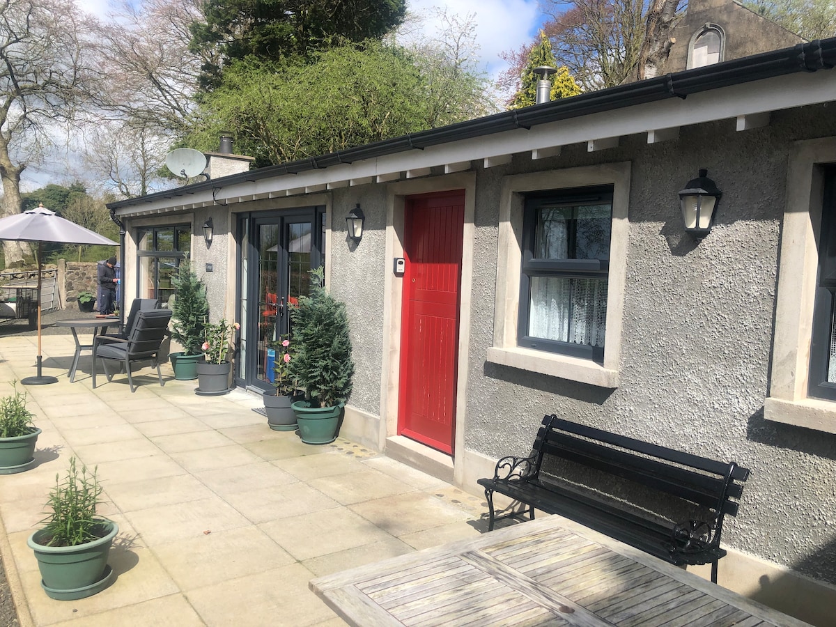 A charming cottage exterior features a bright red door surrounded by large potted plants. The patio area is paved, with a dining table and outdoor furniture, including a shaded umbrella and comfortable seating arranged for relaxation.