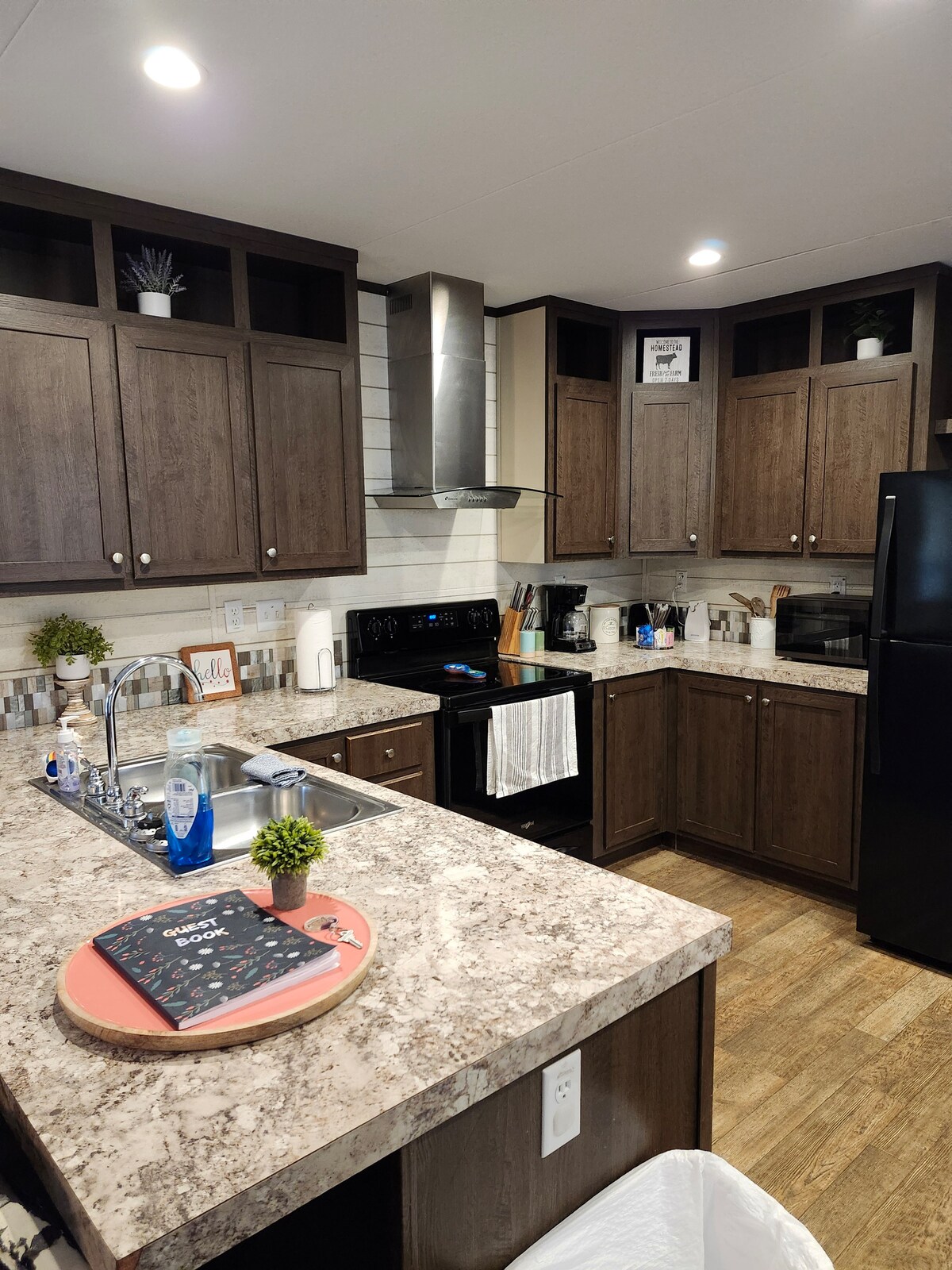 The kitchen features dark wood cabinetry and granite countertops, with an island that includes a sink. Appliances include a black refrigerator and stove. Shelves display plants and utensils, while a tray with items rests on the countertop, contributing to a functional cooking space.