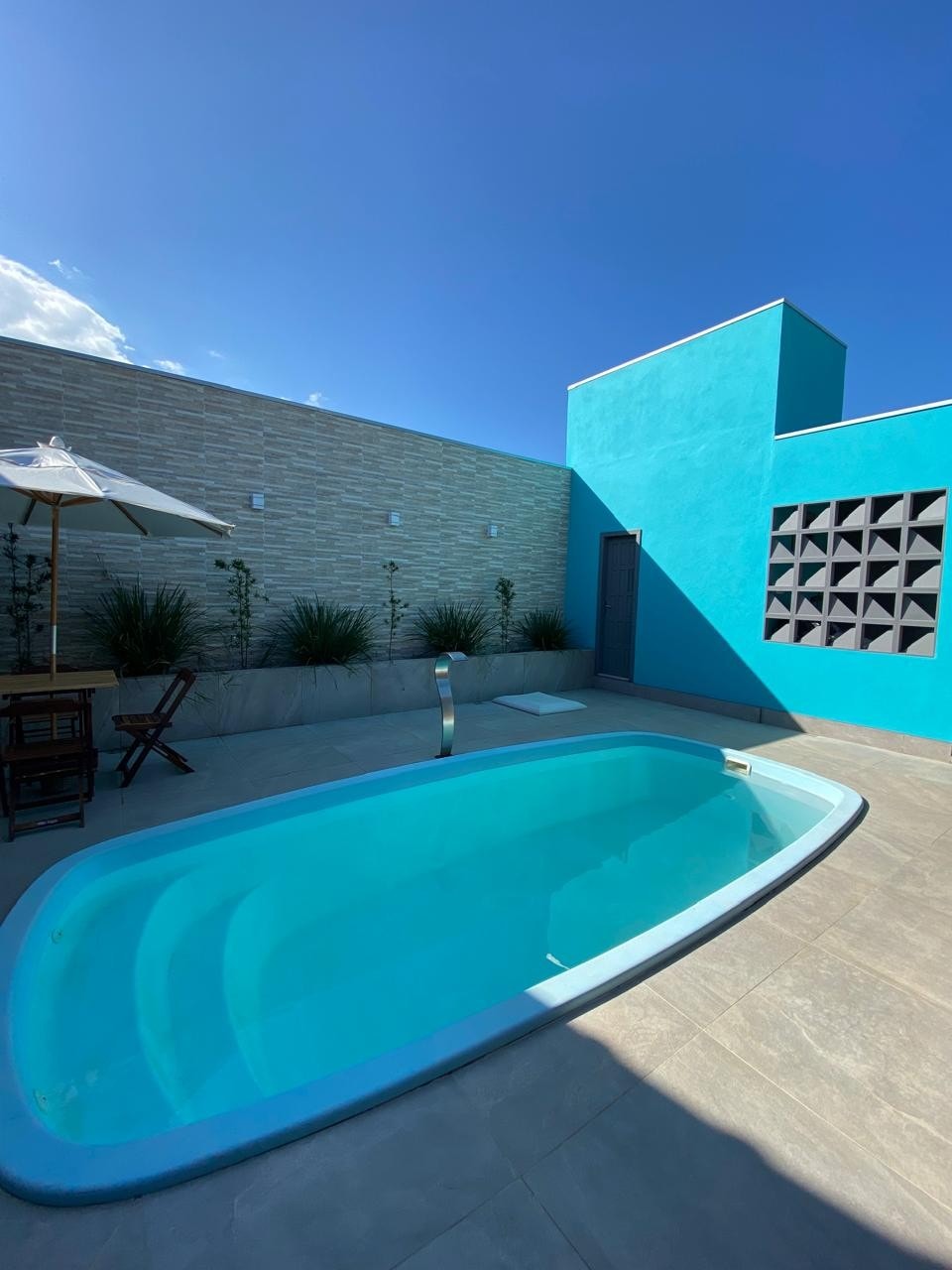 A serene pool area is highlighted with clear, inviting water. Surrounding the pool, a light grey tiled deck is complemented by a stylish umbrella and a wooden table with chairs. A vibrant blue wall adds a pop of color, creating an appealing outdoor space.