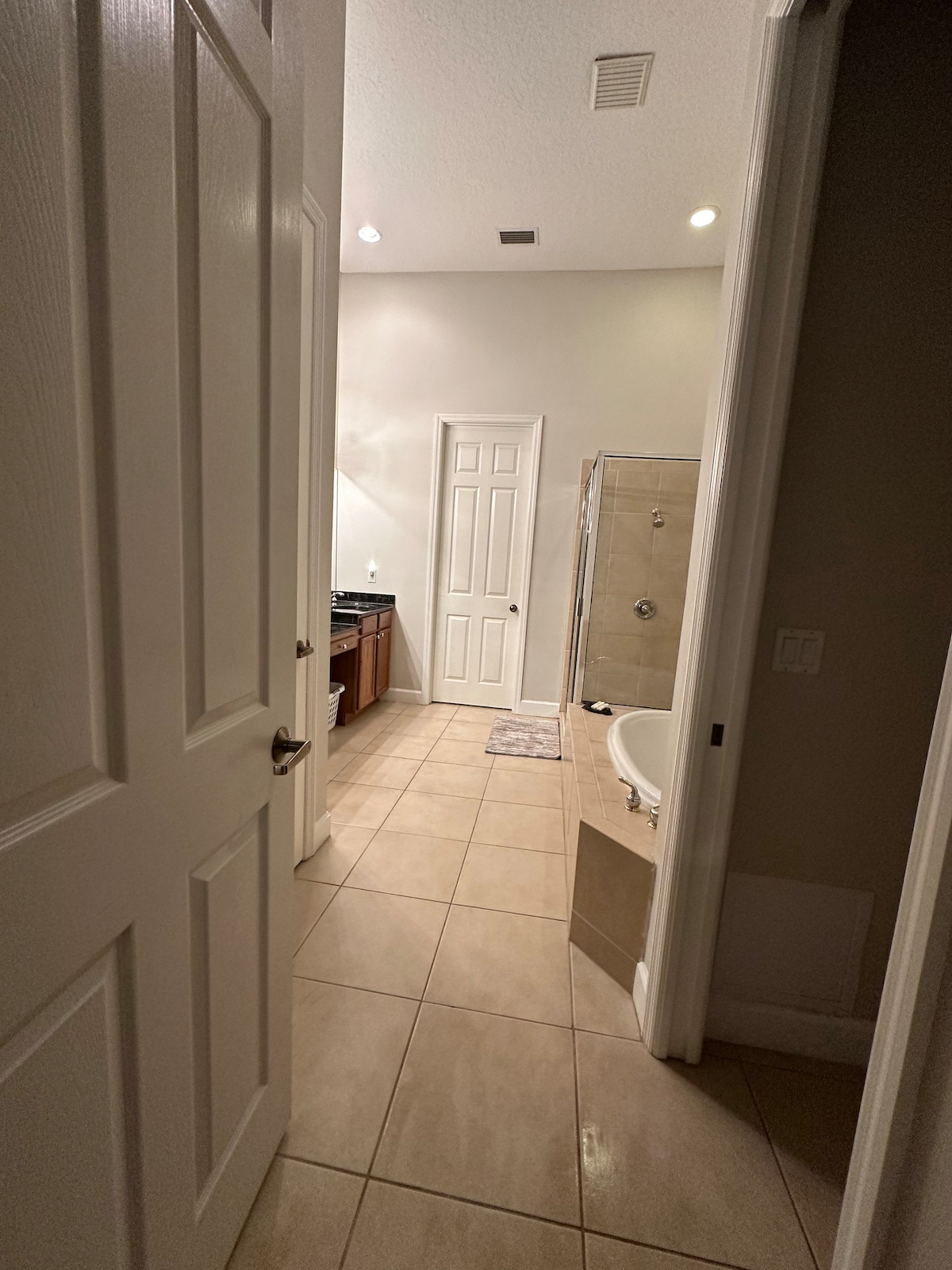 A well-lit bathroom corridor is visible, featuring light-colored tile flooring and warm-toned wooden cabinetry. The doorway leads to a spacious area showcasing a glass shower, a freestanding tub, and a double vanity with sinks, enhancing the overall sense of openness.