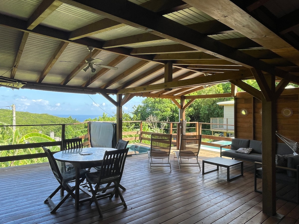 A spacious wooden terrace features a round dining table surrounded by chairs and a lounging area with a sofa. Large sliding doors lead to the private pool, while views of the ocean and greenery are visible in the background.