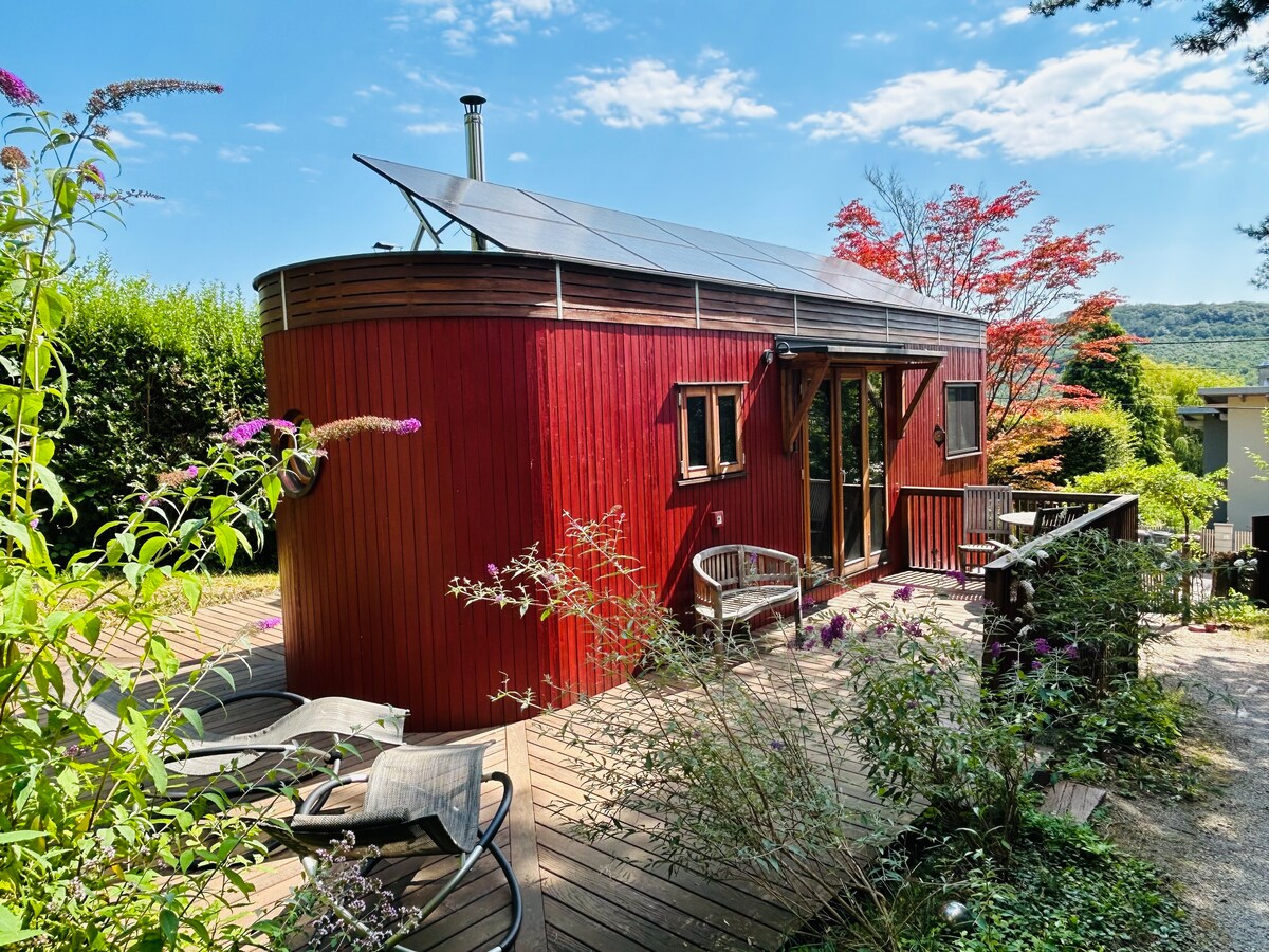 A mobile wooden tiny house showcases a vibrant red exterior with large windows. A solar panel is positioned on the roof. A wooden deck features outdoor seating surrounded by greenery, enhancing the connection with nature.