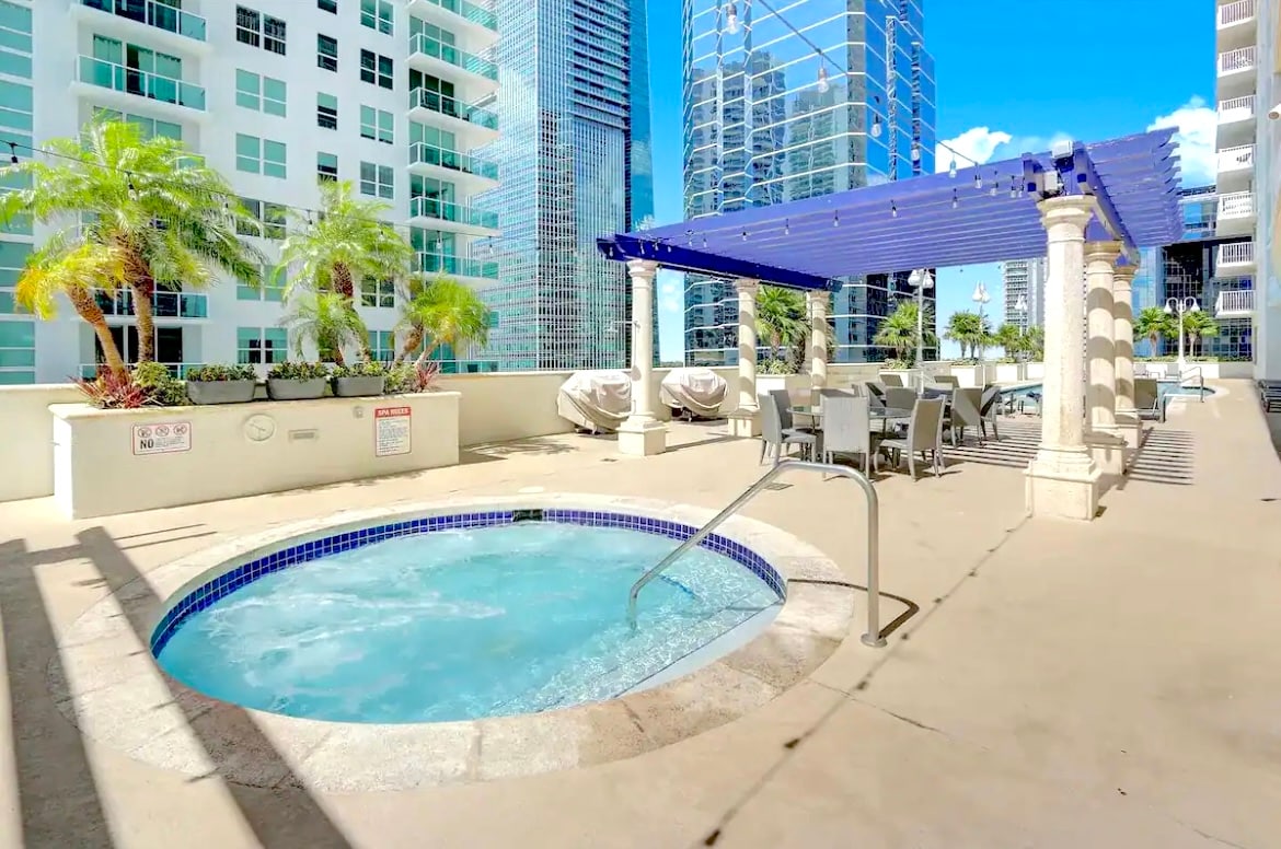 An outdoor relaxation area features a jacuzzi surrounded by a paved deck. Comfortable lounge chairs are positioned nearby under a shaded pergola, with lush palm trees and modern high-rise buildings visible in the background.