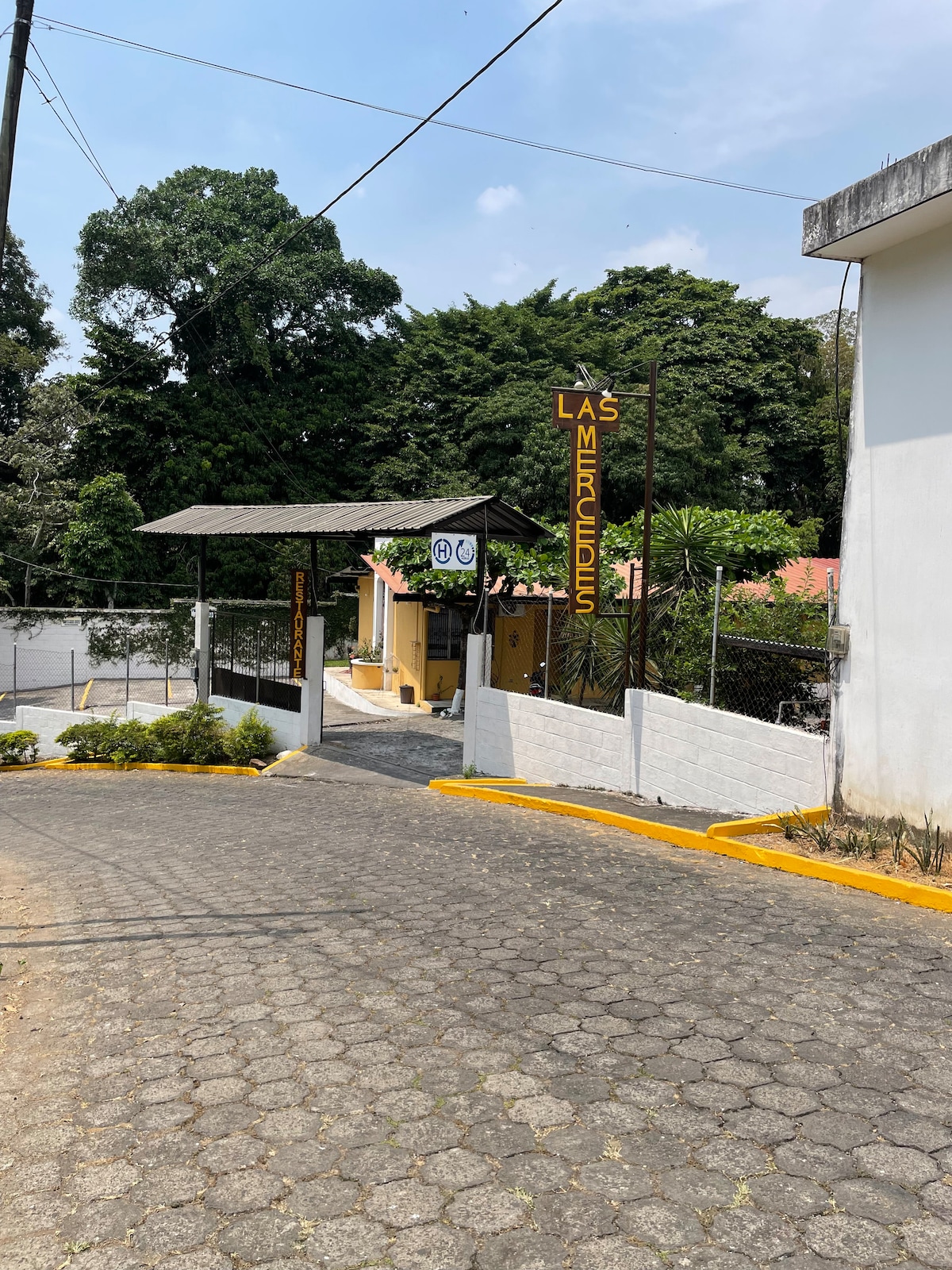 An entranceway to Hotel y Restaurante Las Mercedes is visible, featuring a welcoming sign. The pathway is lined with yellow curbing and bordered by greenery. A canopy offers shaded access to the building, which is partially obscured by trees in the background.