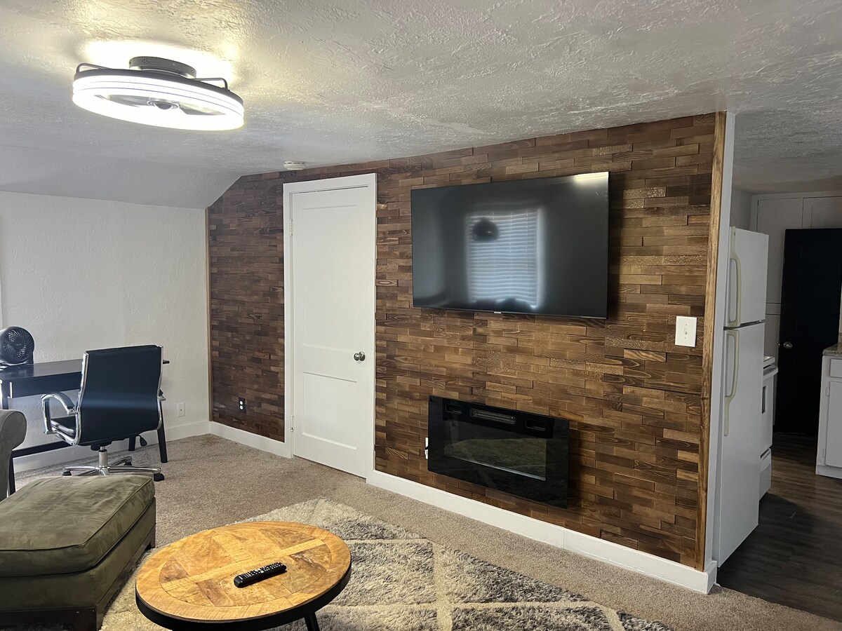 A modern living area features a flat-screen TV mounted on a textured wall, with a cozy electric fireplace beneath. A round coffee table sits in the center, accompanied by a green armchair and a workspace with a desk. A white door leads to a separate space.