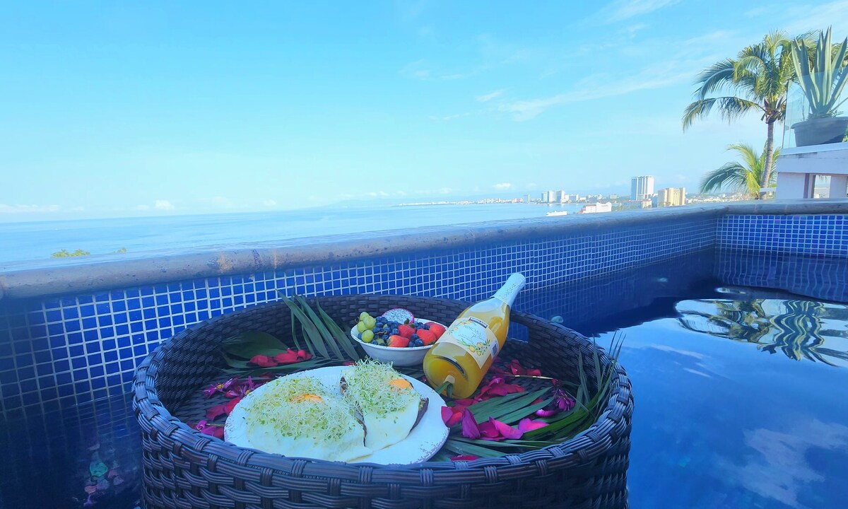 A wicker basket is positioned at the edge of the private pool, filled with a fresh meal garnished with herbs and complemented by a bottle of beverage. The panoramic ocean view stretches into the distance, with the coastline visible under a clear blue sky.