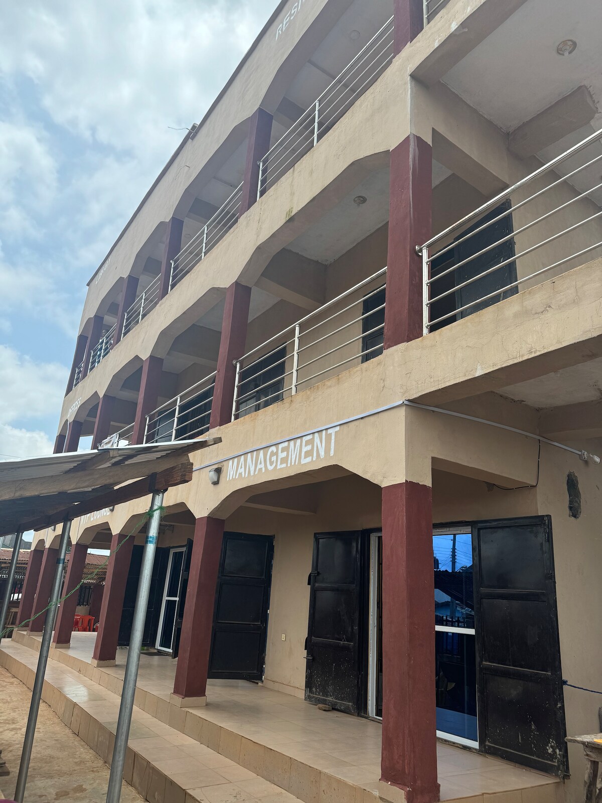 A three-story building is depicted, featuring a light-colored facade with dark accent columns. The entrance displays double black doors, framed by a covered terrace. Balconies with railings line the upper floors, and the word 'MANAGEMENT' is prominently displayed above the entrance.