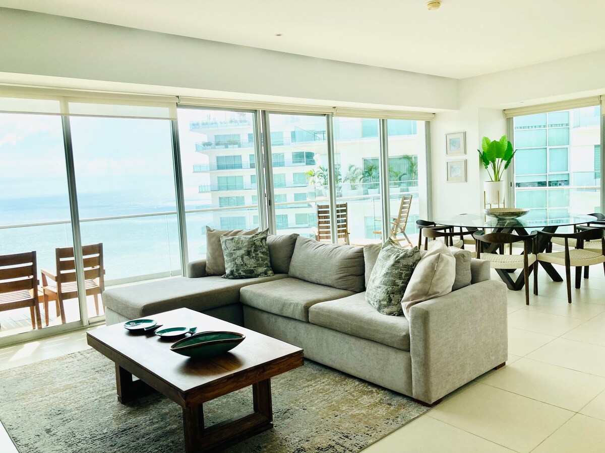 A spacious living area is presented, featuring a comfortable grey sectional sofa paired with accent pillows. A wooden coffee table sits in front, with decorative items arranged atop. Nearby, dining tables and chairs are visible, framed by large windows offering panoramic views of the ocean.