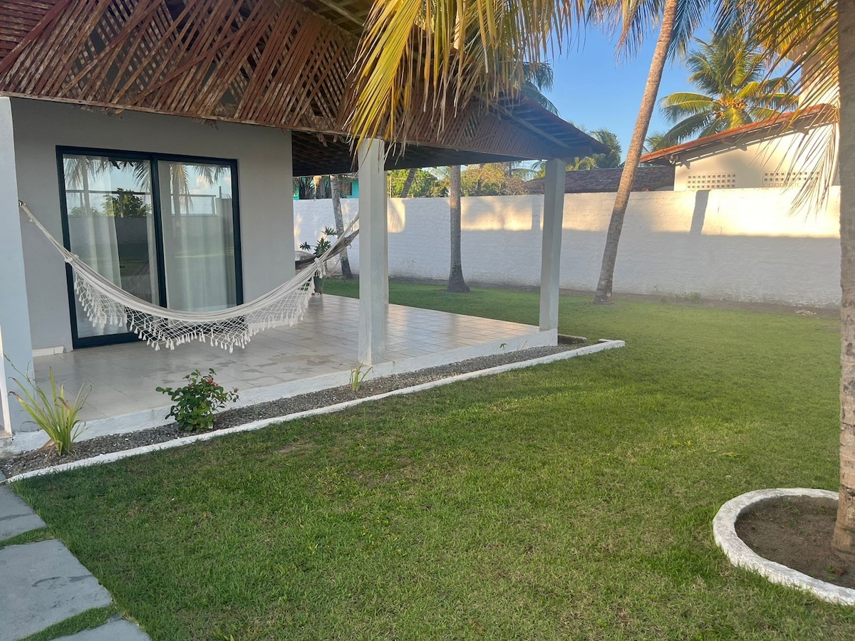 A shaded terrace features a hammock suspended between two posts, surrounded by well-maintained grass. Palm trees provide a tropical ambiance, while the nearby walls offer privacy. Large glass doors lead into the interior space, enhancing the connection with the outdoor environment.