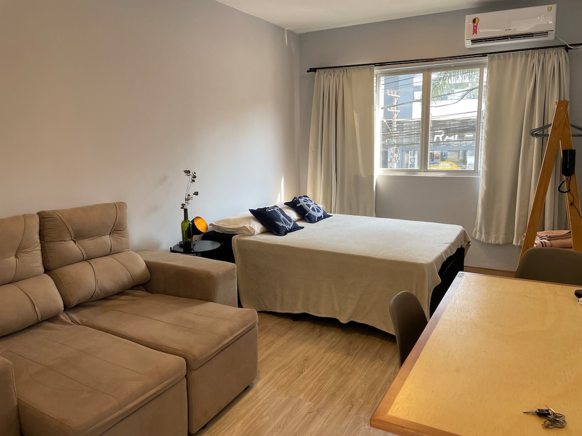 A studio apartment is presented with a cozy seating area featuring a retractable sofa. A spacious bed is outfitted with neutral bedding. Natural light fills the room through a window, accentuated by light curtains. A wooden table is positioned in the corner, complementing the decor.