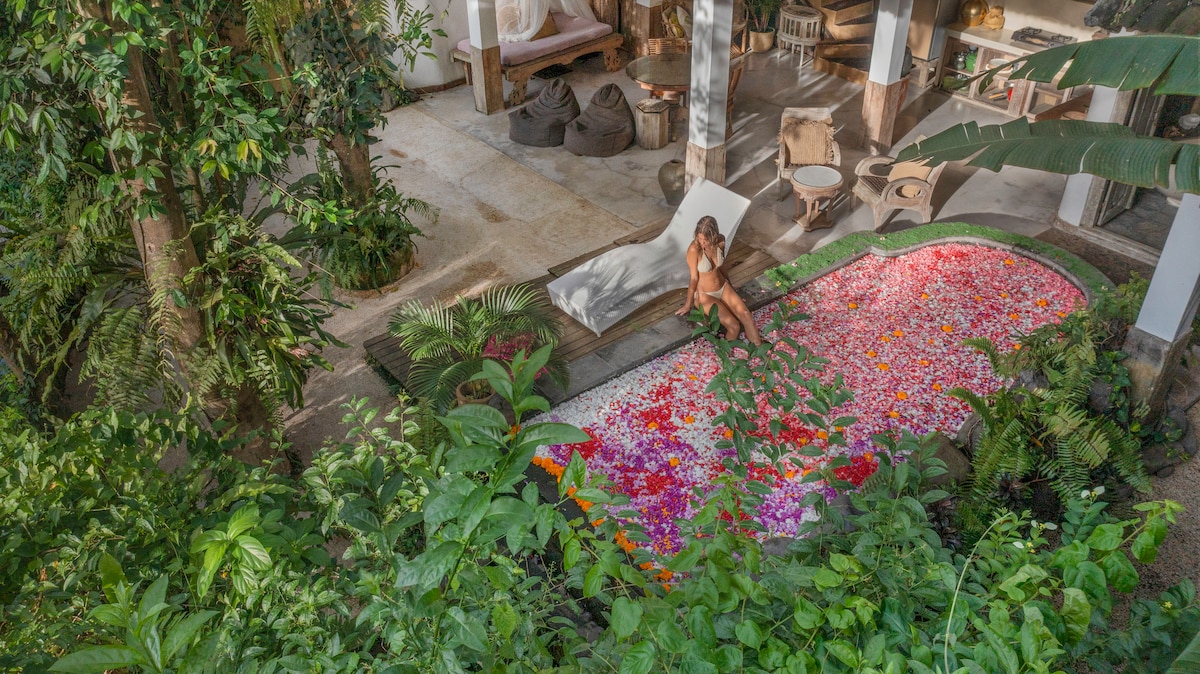 A bright outdoor space is framed by lush greenery, featuring a colorful floral arrangement floating in a private pool. A comfortable lounging area is visible nearby, surrounded by natural textures and tropical plants, creating a serene and inviting atmosphere.