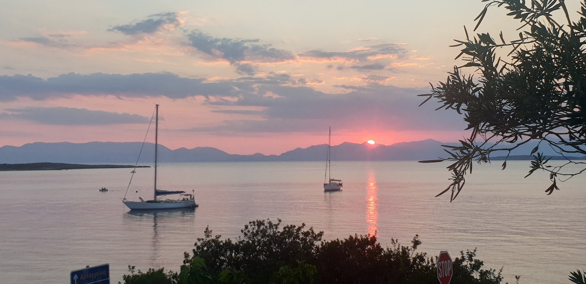 A serene seascape captures a sunset view with boats anchored in calm waters. Soft hues of pink and purple blend in the sky, reflecting on the surface. Silhouettes of distant mountains are visible, framed by lush greenery in the foreground.