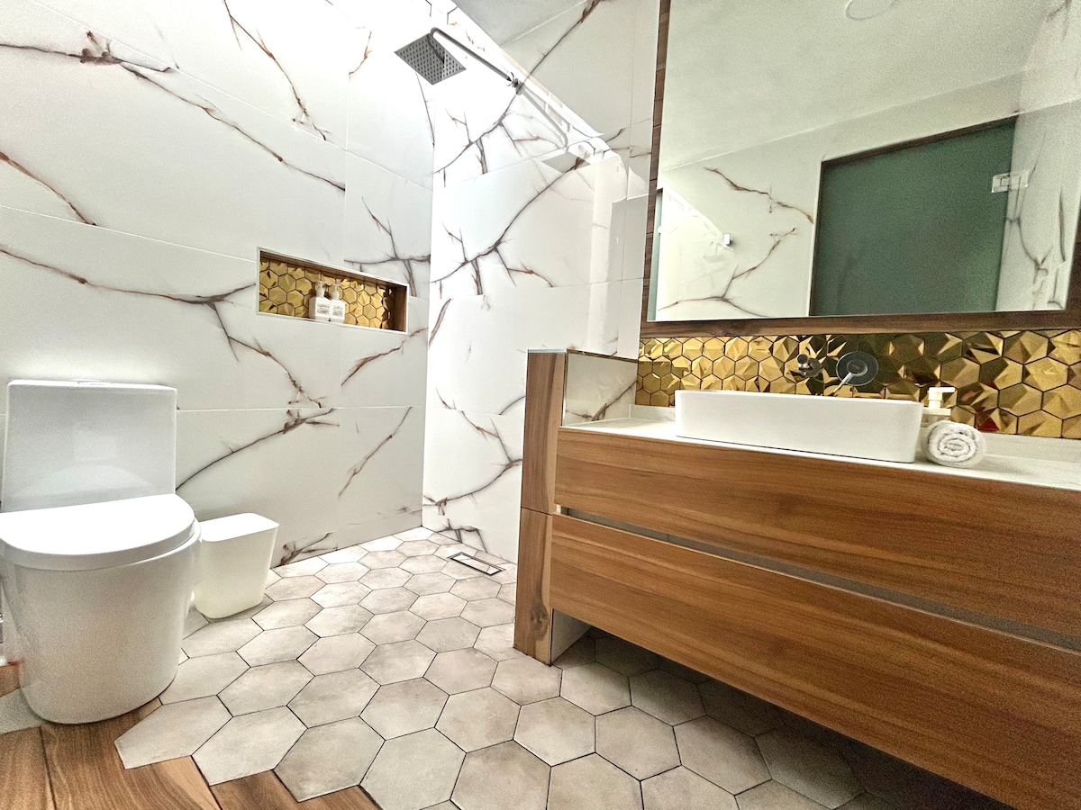 A modern bathroom features a stylish wooden vanity with a vessel sink and gold-accented backsplash. The space is adorned with hexagonal tiles on the floor and is illuminated by natural light from an overhead fixture. A large mirror enhances the room's brightness.