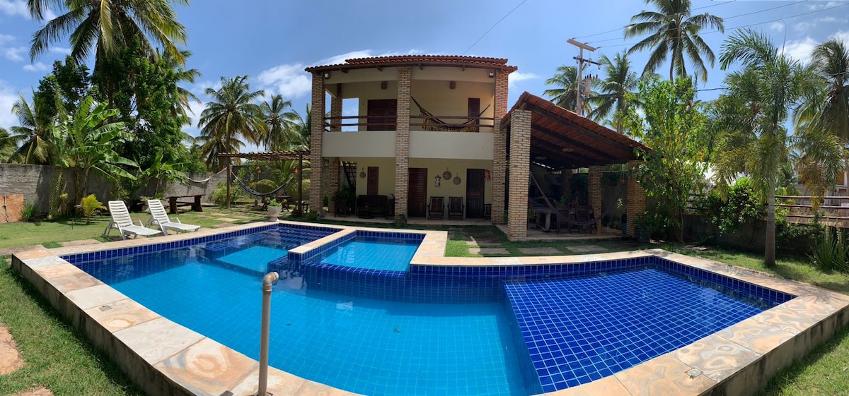 A two-story house is set in a lush tropical landscape, featuring a well-maintained outdoor area. Two inviting swimming pools share a stone patio. Sun loungers are positioned near the water, while palm trees gently sway in the background beneath a clear blue sky.