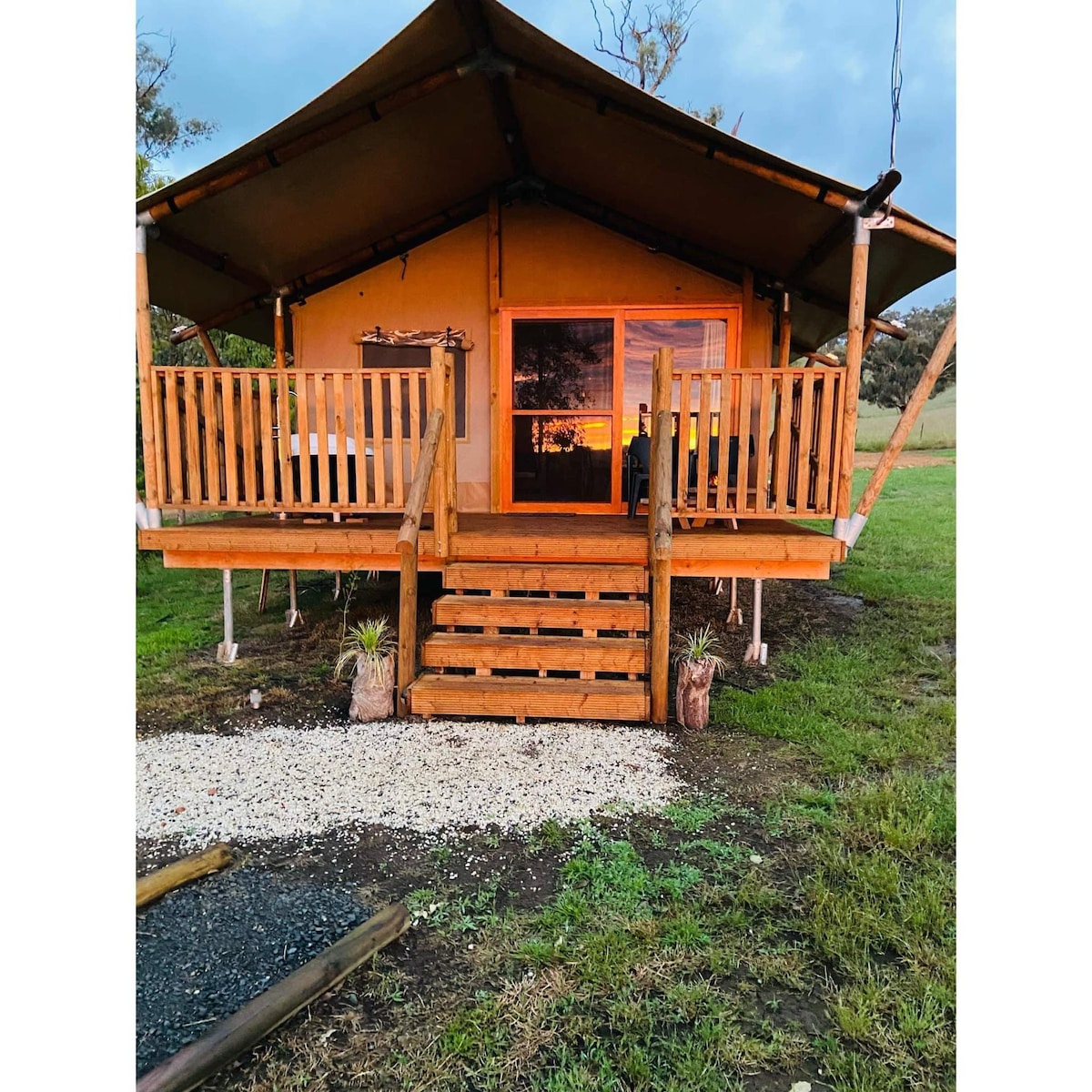 A raised glamping tent features a welcoming wooden deck with sturdy railings and steps leading up to the entrance. Large windows provide natural light, while the sunset casts warm hues across the exterior, highlighting the tent's unique setup surrounded by open grassland.