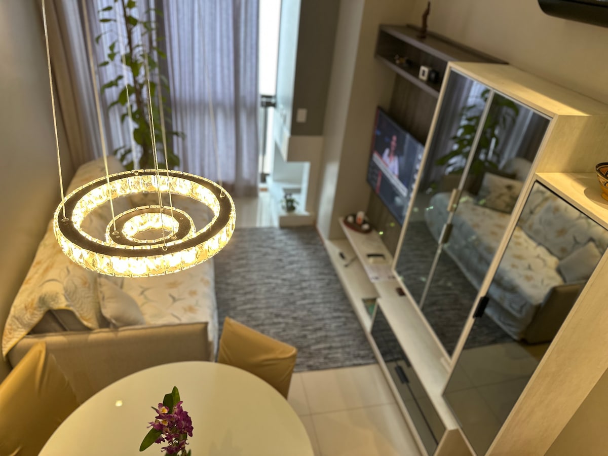 A well-lit living space features a round dining table surrounded by soft-colored chairs. A modern chandelier hangs above the table, illuminating the area. A sleek entertainment unit with a Smart TV is positioned against the wall, while a comfortable sofa is seen in the background, enhancing the inviting atmosphere.