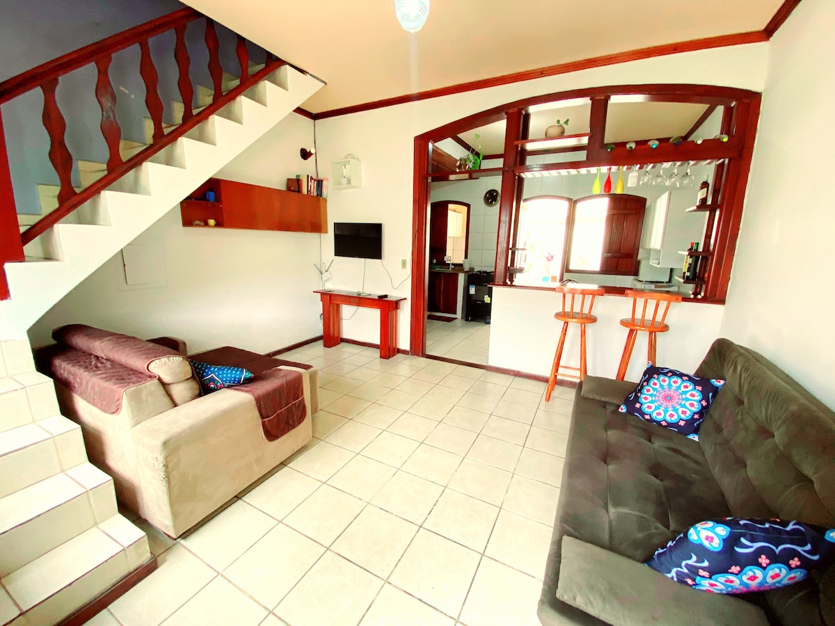 A well-organized living area features two comfortable sofas and a wooden bar with two stools. Natural light pours in through multiple windows, illuminating the tiled floor. Stairs lead to the upper level, while a small entertainment unit is positioned against the wall.