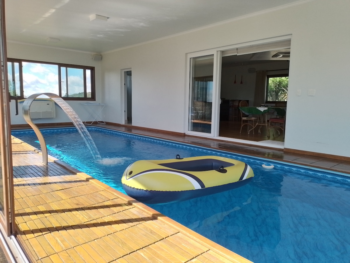 An indoor pool is visible, featuring a bright yellow inflatable float resting on the water. A waterfall feature flows gently into the pool, and large glass doors provide access to the outdoor area, allowing natural light to fill the space.