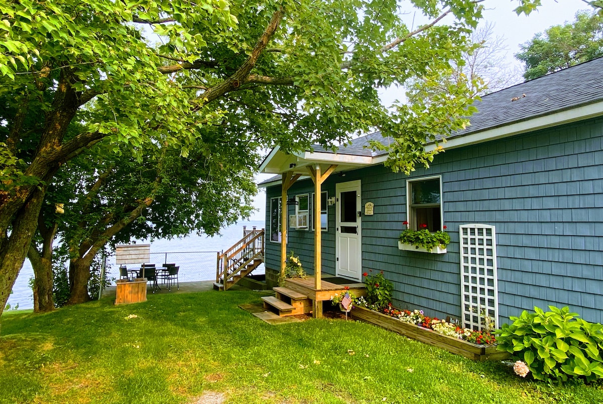 The entrance of Bluebird Cottage is framed by lush greenery and vibrant flower beds. Sturdy steps lead up to a welcoming door, while a patio set is visible nearby, offering views of Lake Champlain. The serene surroundings create a peaceful outdoor environment.