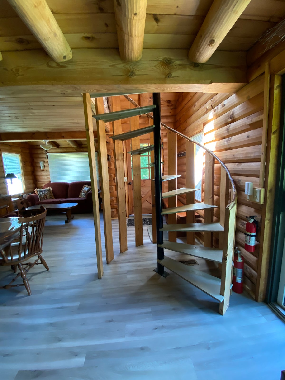 A spiral staircase crafted from wood is featured, leading to a sleeping loft. The interior is finished with natural wood paneling, complemented by light-colored flooring. A cozy living area, partially visible, includes a sofa and dining space, illuminated by natural light from nearby windows.