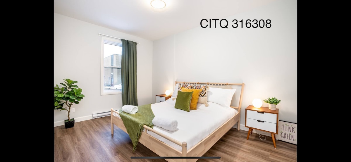 A serene bedroom features a spacious bed adorned with fresh linens and decorative pillows. Natural light filters through a large window with green curtains, complementing the minimalist decor. A potted plant adds a touch of nature, while nightstands with soft lighting provide a sense of warmth.