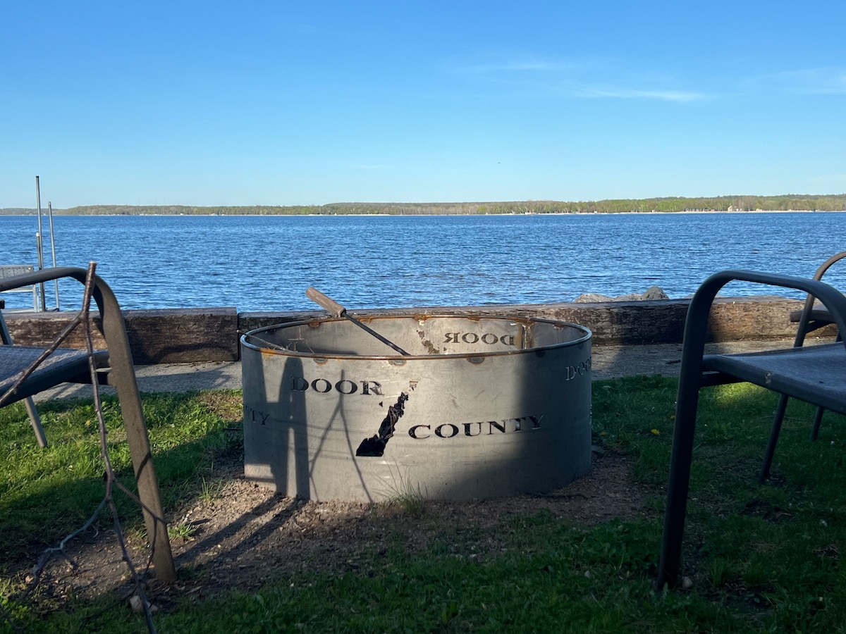 A firepit is set on a grassy edge overlooking the water. Metal walls are engraved with 'Door County.' Nearby, a few chairs are positioned for seating, while the lake stretches into the distance under a clear sky.