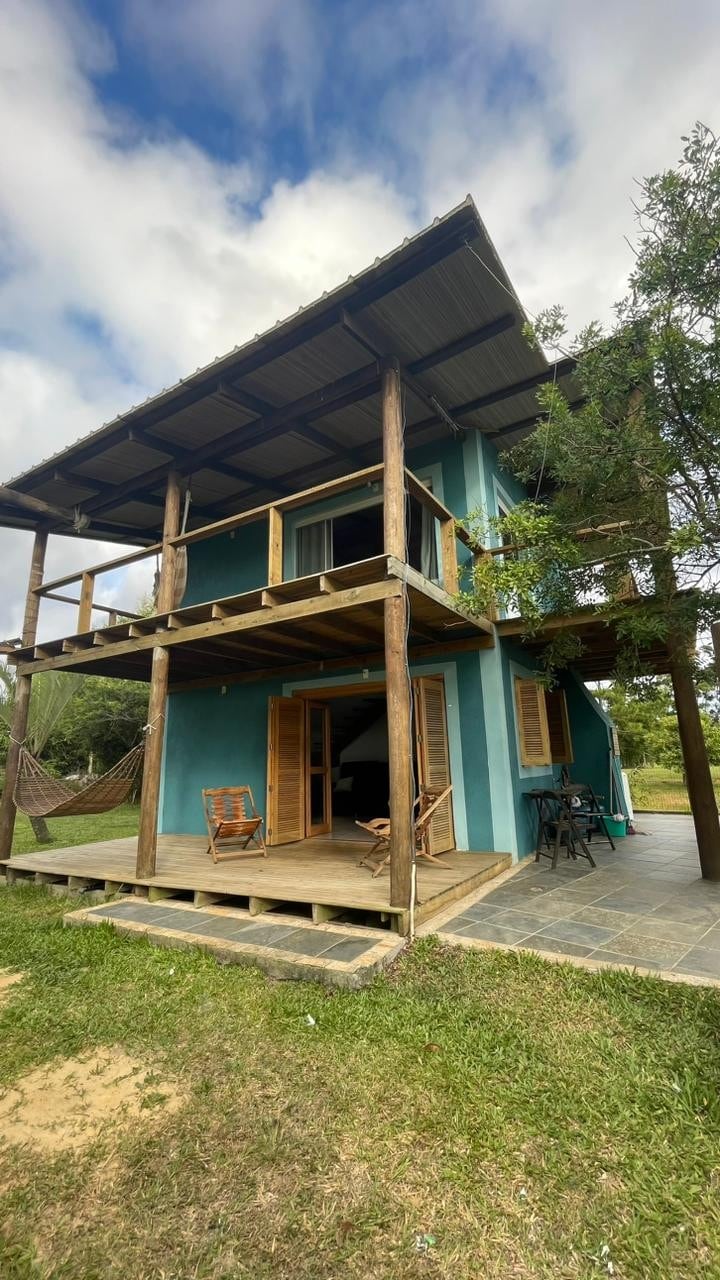 A two-story house is visible with a vibrant blue exterior. A spacious wooden deck is adorned with two comfortable chairs and a hammock. Large doors open to the exterior, inviting natural light. Lush greenery surrounds the property, enhancing the tranquil atmosphere.
