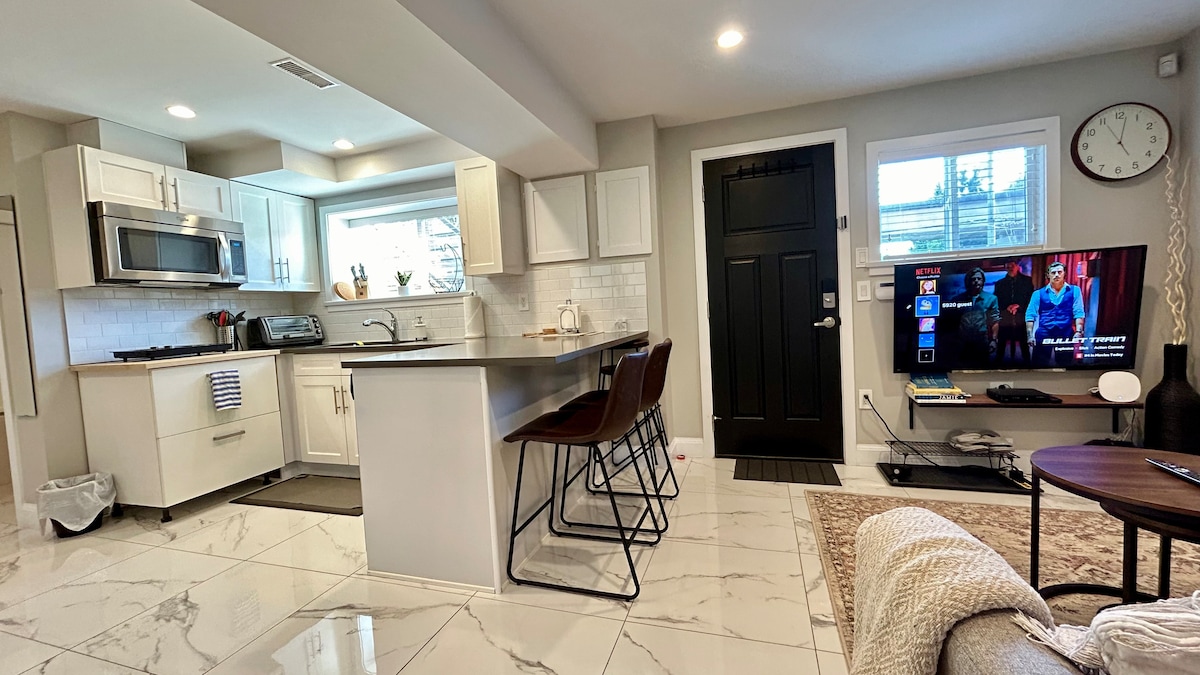 A modern kitchen area is showcased with sleek white cabinetry and high-end appliances. A spacious countertop provides seating for three on stylish bar stools. Nearby, a living area features a flat-screen TV and a cozy rug, with natural light filtering through a window.