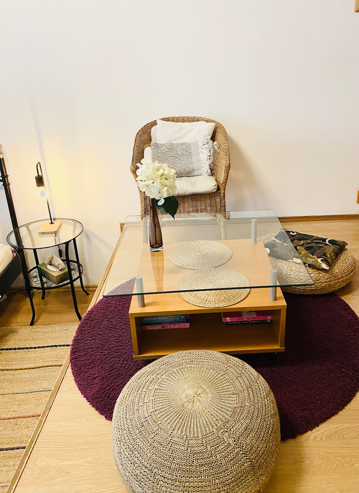 A cozy living space features a glass-top coffee table centered on a round maroon rug. Several textured poufs provide seating around the table. A rattan chair is positioned nearby, adorned with decorative cushions. A small side table holds a lamp and a vase with flowers.