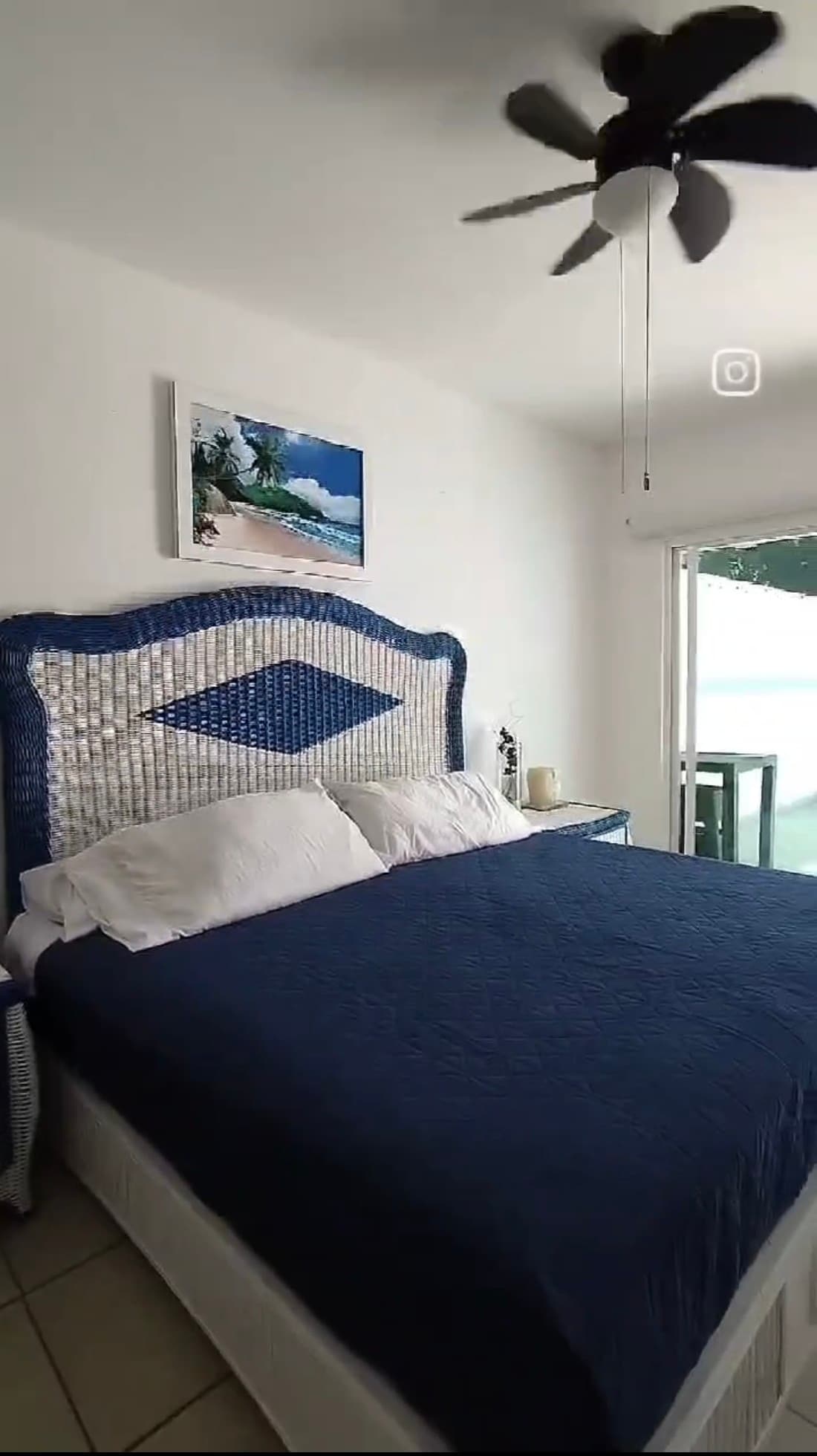 The bedroom showcases a spacious bed featuring a blue quilted cover and white pillows. A fan hangs overhead, and a large window opens to an outdoor area, allowing natural light to brighten the room. A painting of a beach scene decorates the wall.