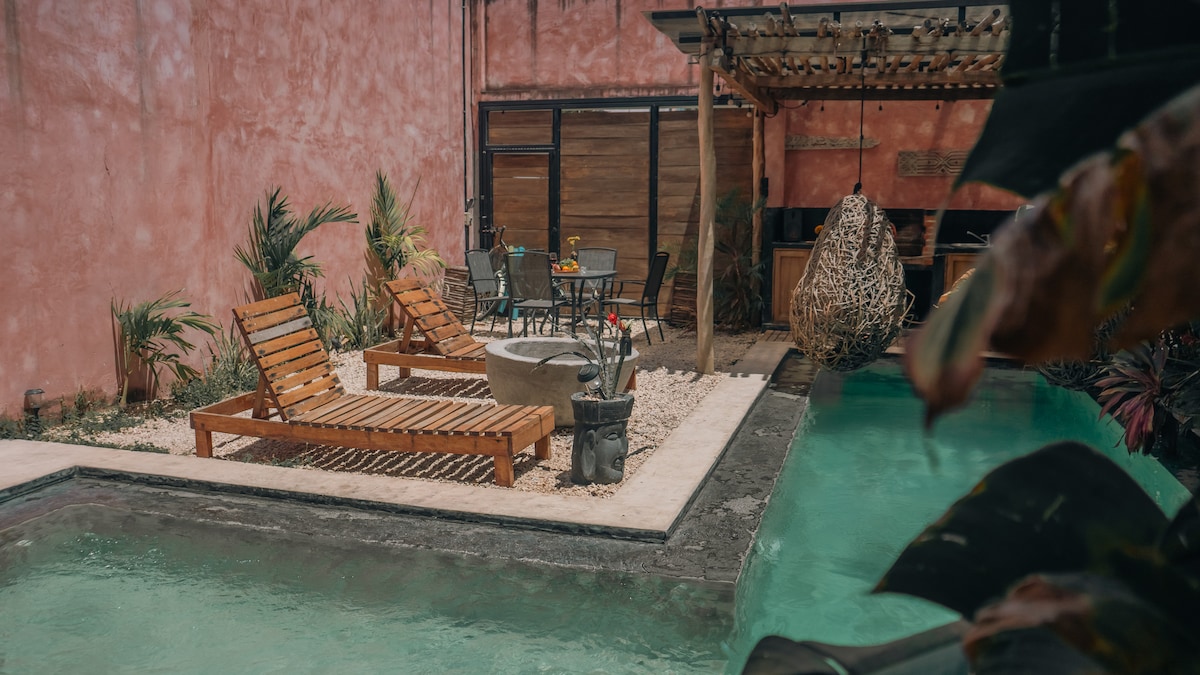 A tranquil outdoor area features two wooden lounge chairs positioned near a circular stone table, surrounded by lush plants. A cozy hanging chair is visible under a wooden pergola, enhancing the inviting atmosphere of the small swimming pool in the backdrop.