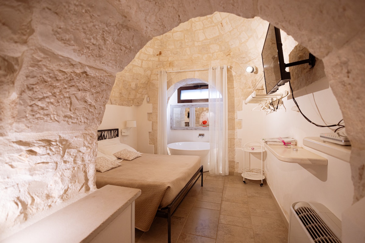 The room features a curved stone ceiling and soft lighting, enhancing the cozy atmosphere. A spacious bed is positioned near a modern bathtub, while a wall-mounted television is visible above a small shelf. Natural light enters through a window adorned with sheer curtains.