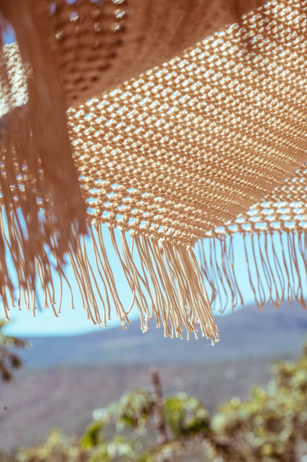 A woven textile with intricate patterns hangs overhead, showcasing decorative fringes. The background features a blurred view of lush greenery and mountain peaks under a clear blue sky, creating a serene outdoor atmosphere.