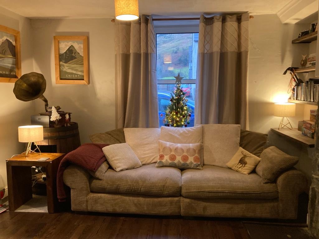 A comfortable lounge area features a large sofa adorned with patterned cushions. A small table beside it holds a lamp. A window allows natural light in, framed by light curtains. A decorative Christmas tree stands in the corner, adding a festive touch to the room.