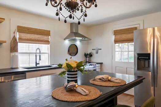 A modern kitchen is highlighted by a black chandelier and bamboo window shades. A large island features two place settings with napkins and decorative woven placemats. Stainless steel appliances are visible, along with a window overlooking the exterior.