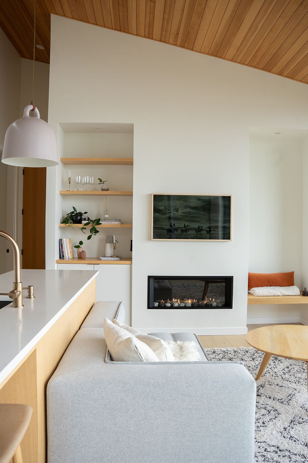 The open-concept living area features a modern sofa facing a sleek gas fireplace and a wall-mounted television. Natural wood accents complement the light-colored walls, while shelves display plants and books. A round wooden table, positioned nearby, adds a practical touch to the relaxed space.