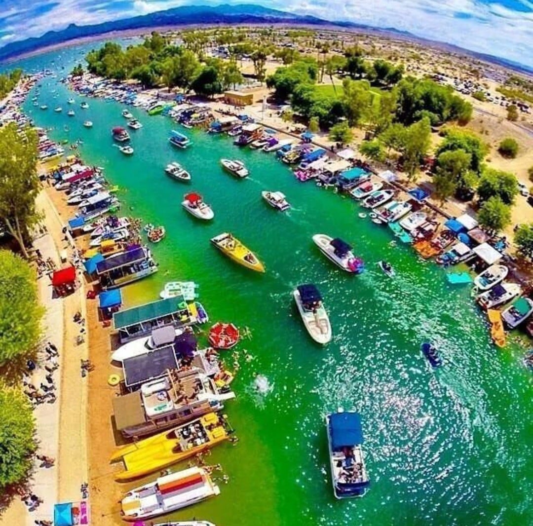 An aerial view captures a busy waterway lined with boats and watercraft. Lush greenery borders the turquoise waters, which are filled with recreational vehicles. Sandy paths lead to various docking areas, surrounded by shaded spots featuring umbrellas and canopies for relaxation.