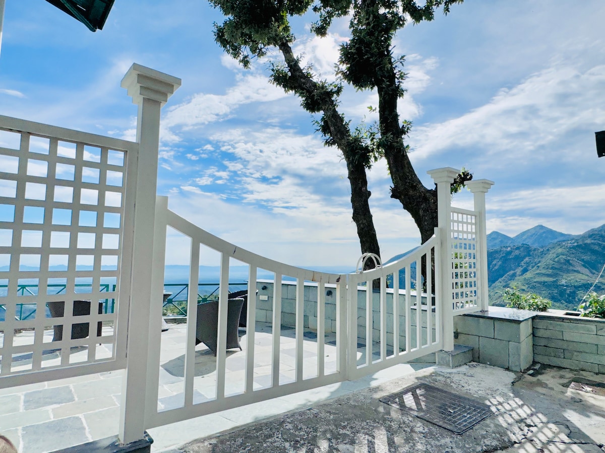 A gated entrance is framed by white lattice work, leading to a serene outdoor space. The view beyond features a sweeping landscape of mountains under a clear blue sky, with the sea visible in the distance. Stone flooring provides a sturdy path to enjoy the surroundings.