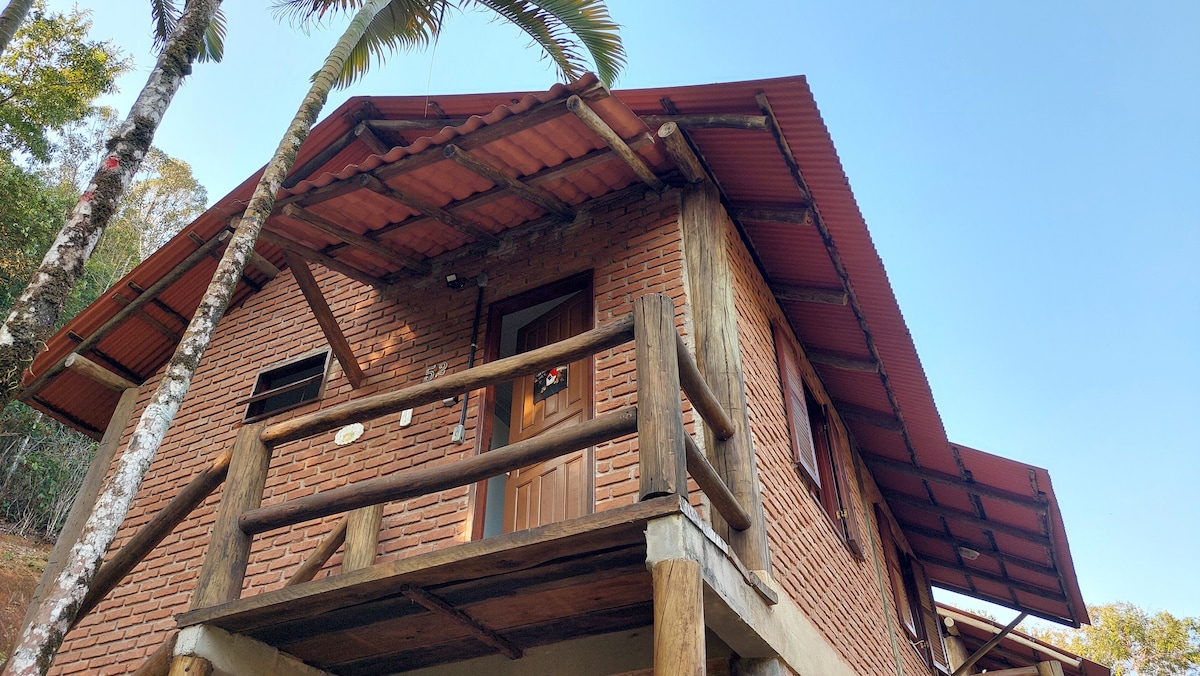 The exterior of the chalet features rustic brick walls and a red-tiled roof. A wooden balcony provides a space to enjoy the surroundings, while palm trees are visible nearby. The entrance door is centrally located, framed by large wooden beams.